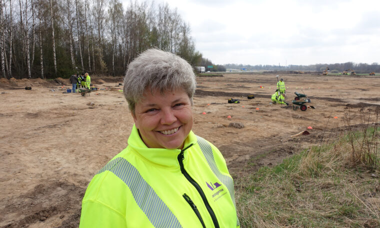 Kvinna i gula arbetskläder framför utgrävning på slätt