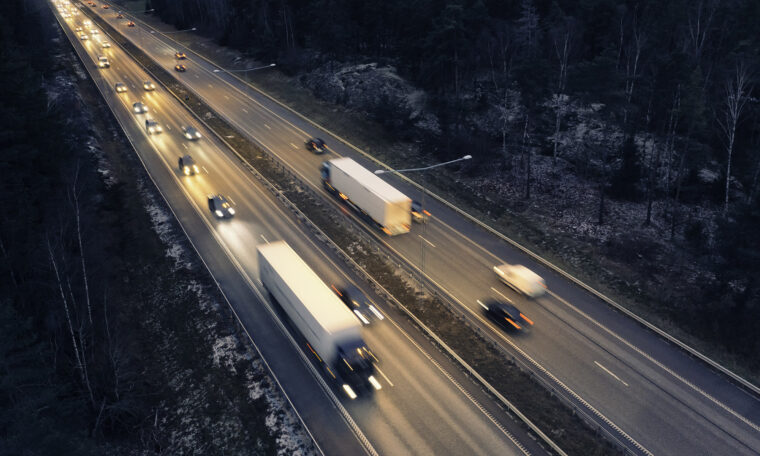 Motorväg genom en skog med trafik efter solnedgången, med fläckar av tidig snö på marken.