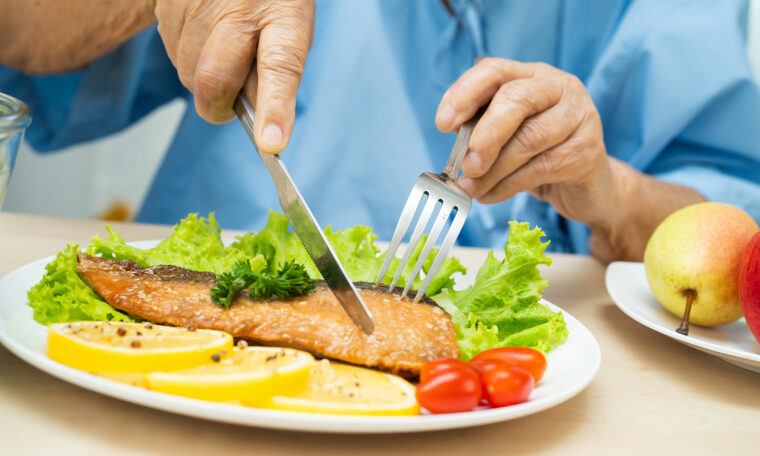 Lax och grönsaker på en tallrik. En äldre persons händer håller i besticken