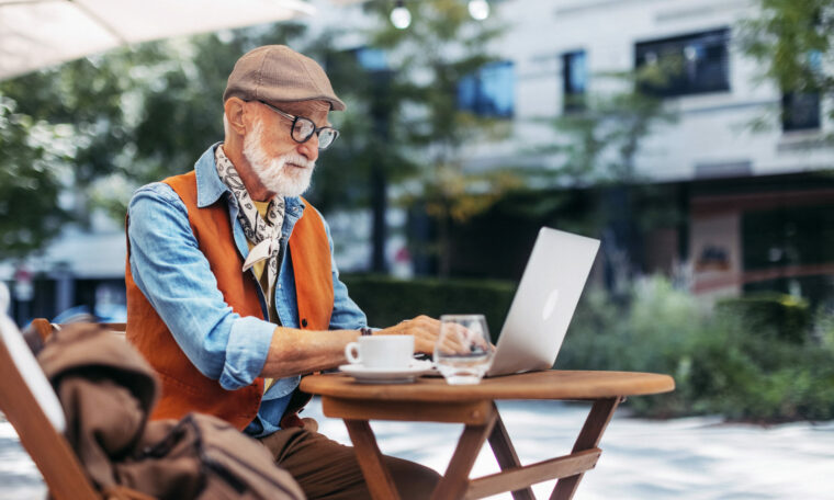 Äldre man sitter på en uteservering och skriver på en laptop.