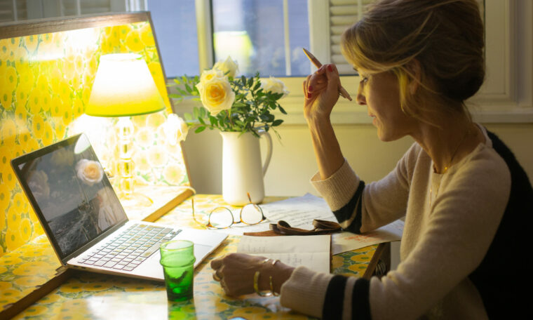 En kvinna sitter vid sin dator och skriver i ett anteckningsblock.
