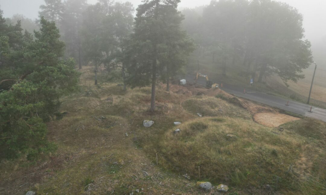 Utgrävning i kuperat landskap med tallar intill en landsväg.
