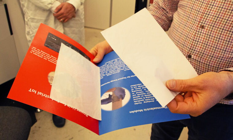 En person håller upp flera tunna, böjliga ark av material framför röda och blå presentationsblad.